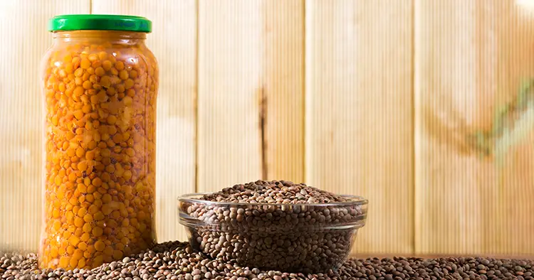 Front view of raw lentil grains in glass bowl and canned lentils in jar on wooden background