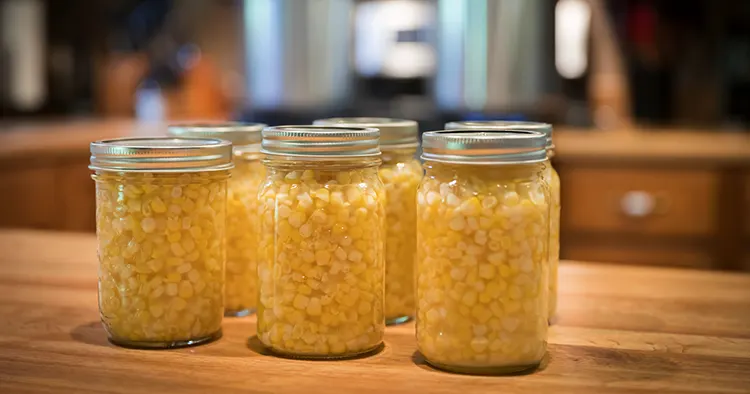 Pint jars of corn sitting on the counter ready to go in pressure canners that are sitting on the stove in the background.