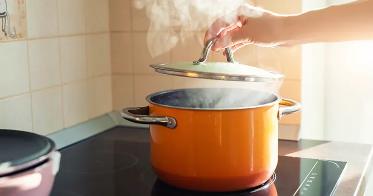 Female hand open lid of enamel steel cooking pan on electric hob with boiling water or soup and scenic vapor steam backlit by warm sunlight at kitchen