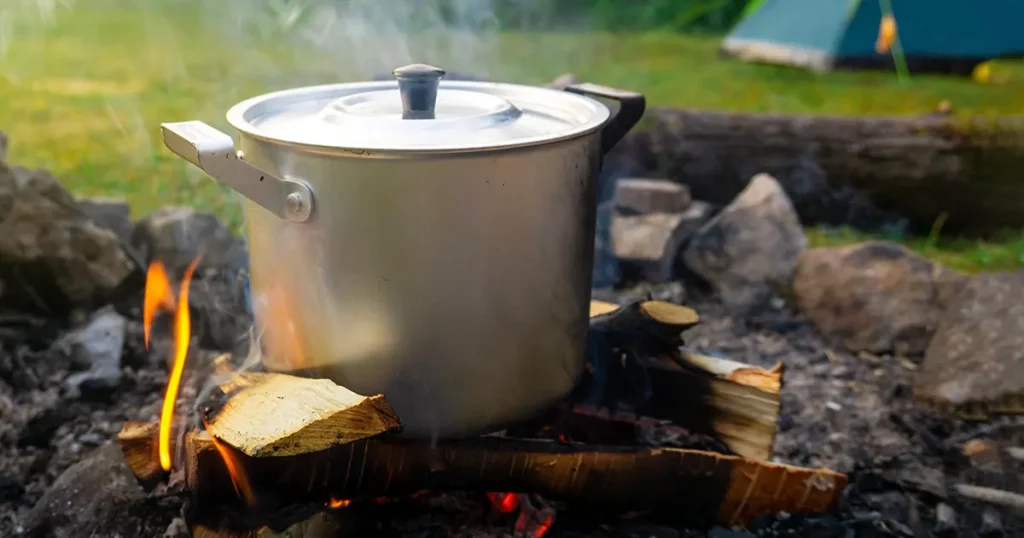 A steaming saucepan stands over an open fire. In the background there is a forest and a tent